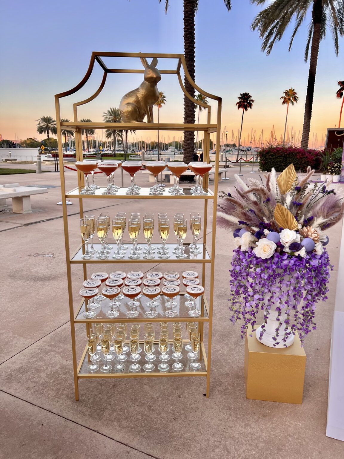 Sophisticated drink display with a variety of cocktails and champagne glasses on a gold shelf, set against a stunning sunset backdrop, ideal for an upscale outdoor event.