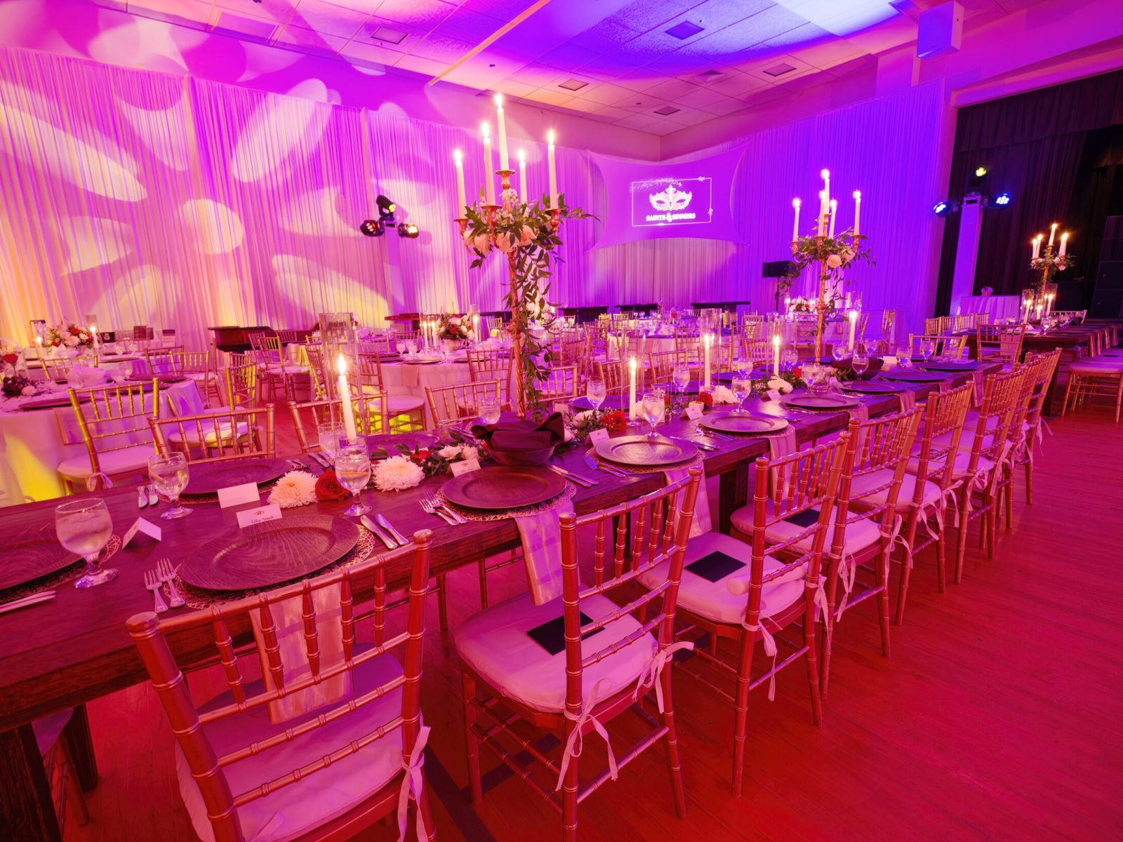 Candlelit banquet setup with long tables, golden chairs, and ambient purple lighting, ideal for a sophisticated gala or wedding reception.