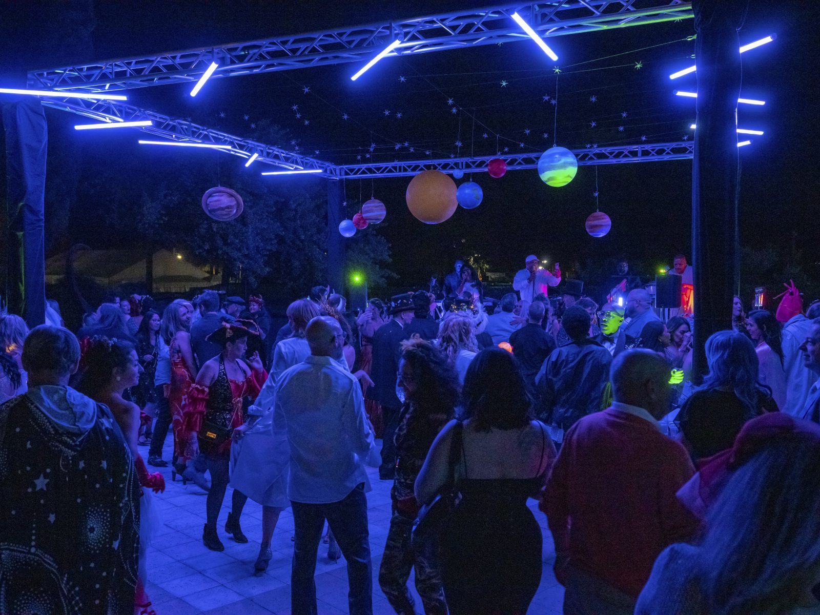 Crowded outdoor night event with a glowing neon canopy and planetary decorations, creating a lively party atmosphere under the Florida sky.