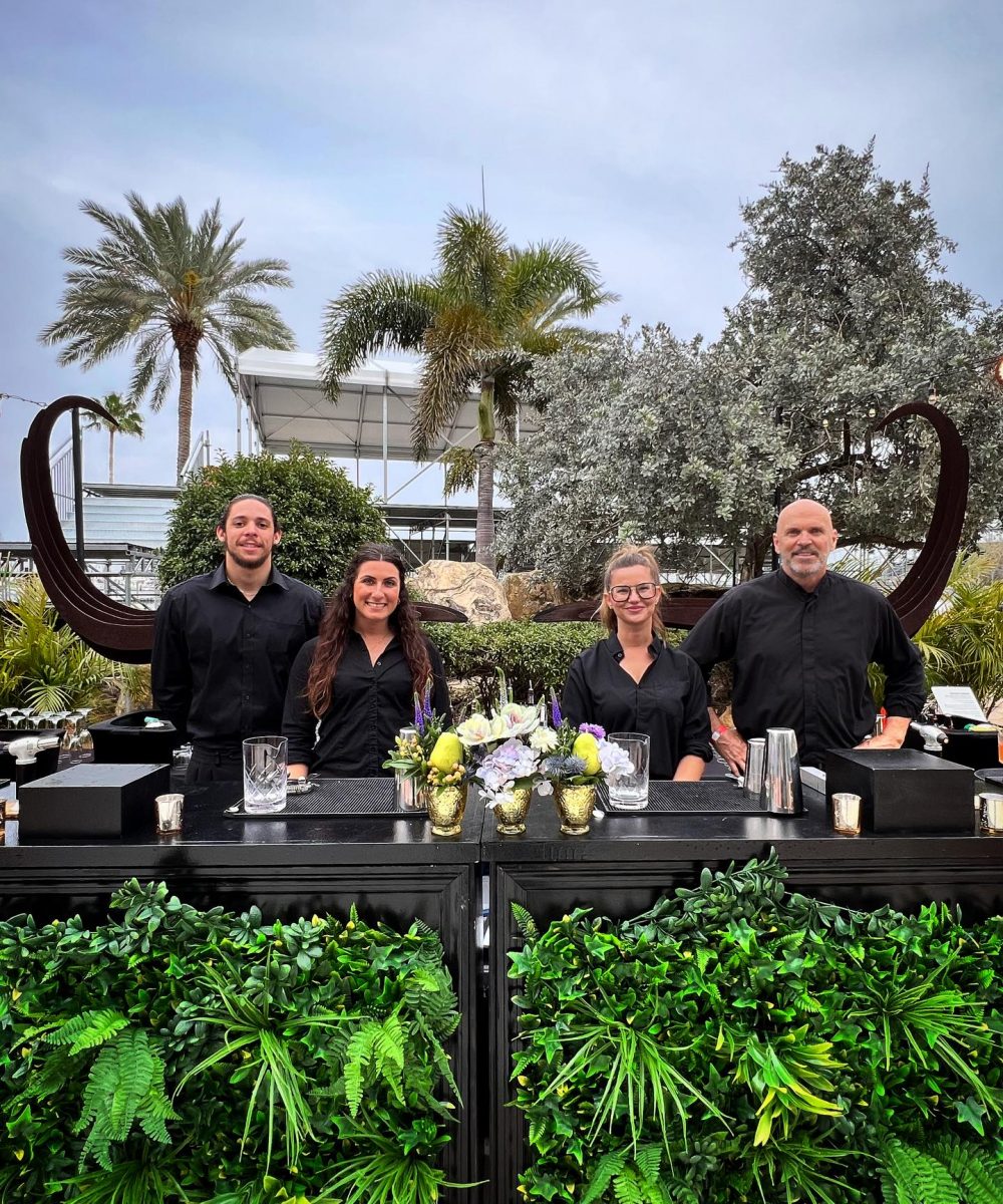 Professional bar staff standing behind a lush greenery-adorned bar, ready to serve at an upscale outdoor event on the Gulf Coast.